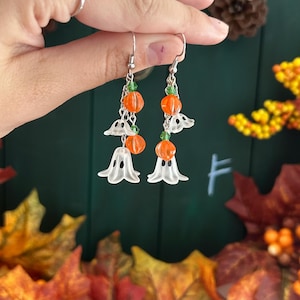 Op de afbeelding: Een paar Halloween-oorbellen. Elke oorbel heeft een zilveren haak, groene kralen, oranje pompoenkralen en een witte spookbedel. De oorbellen worden gepresenteerd tegen een achtergrond van herfstbladeren.