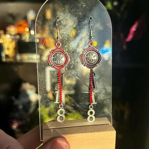 Op de afbeelding: Zilveren oorbellen met een rood en zilver brandweerembleem ontwerp. De oorbellen hebben een bungelende ketting met de nummers "18" onder het embleem. De oorbellen worden tentoongesteld op een heldere acryl standaard.