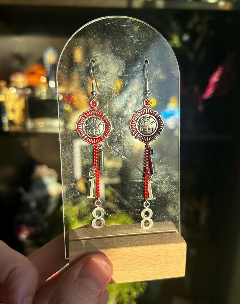 Op de afbeelding: Zilveren oorbellen met een rood en zilver brandweerembleem ontwerp. De oorbellen hebben een bungelende ketting met de nummers "18" onder het embleem. De oorbellen worden tentoongesteld op een heldere acryl standaard.