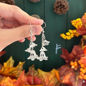 Op de afbeelding: Een paar zilverkleurige oorhangers met doorschijnende witte spookvormige bedels. Elke oorbel heeft een ketting met drie spookbedels. De oorbellen worden omhoog gehouden tegen een achtergrond van herfstbladeren en een donkergroen oppervlak.