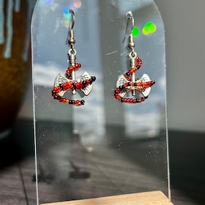 以下が含まれることがあります： 赤と黒のビーズで飾られた銀の斧のイヤリングのペア。イヤリングは透明なアクリル製のディスプレイスタンドに吊り下げられています。