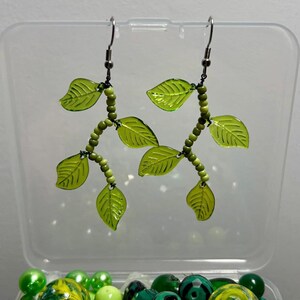 Op de afbeelding: Een paar groene bladeren oorbellen met zilveren haken. De oorbellen zijn gemaakt van groene plastic bladeren en kleine groene kralen.