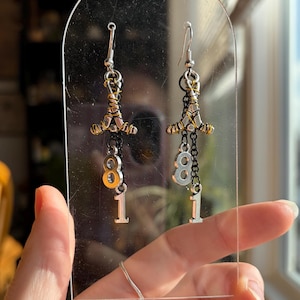 May include: A pair of dangle earrings with silver hooks. Each earring features a silver and gold-toned metal design with a chain holding a silver number 8 and a silver number 1 charm. The earrings are displayed against a clear background.
