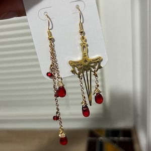 May include: A pair of gold earrings with red glass beads. One earring features a sword design with a star at the top. The other earring has a chain with multiple red beads.