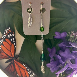 May include: A pair of earrings featuring a silver feather charm and a dangling chain with green crystal beads. The earrings are shown on a white card against a green backdrop.