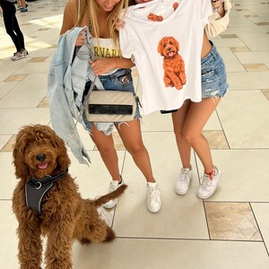 May include: Two young women are holding a white t-shirt with a brown dog illustration. The dog is wearing a red collar and has a happy expression. The t-shirt has the words "Harvard" printed on it. The women are standing in a shopping mall with a brown dog on a leash in front of them.