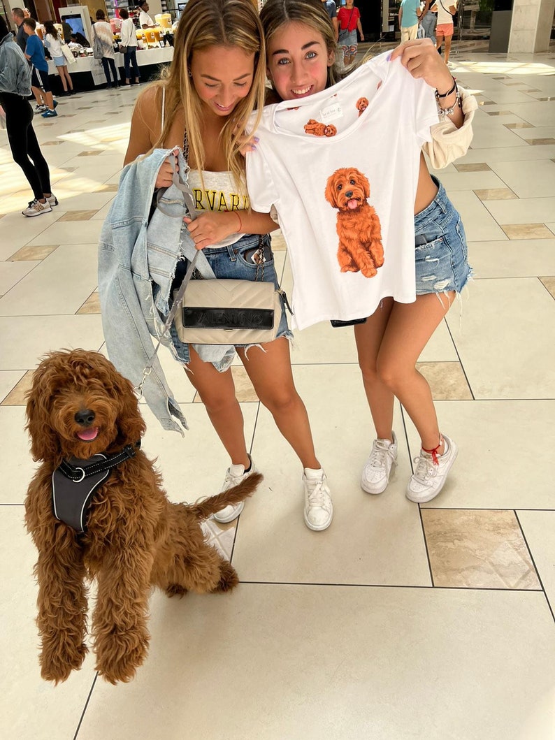 May include: Two young women are holding a white t-shirt with a brown dog illustration. The dog is wearing a red collar and has a happy expression. The t-shirt has the words "Harvard" printed on it. The women are standing in a shopping mall with a brown dog on a leash in front of them.