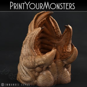 May include: A 3D printed model of a monster's head with an open mouth, revealing rows of teeth. The model is made of a light brown material and has a textured surface. The model is titled "PRINT YOUR MONSTERS".