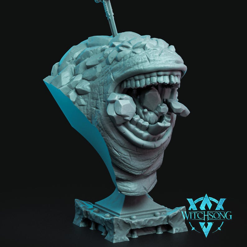 May include: A gray, stone-like sculpture of a creature with a large, open mouth filled with rocks. The sculpture is on a base with the text "WITCHSONG" on it.