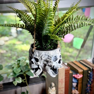 May include: A hanging planter shaped like a cluster of three human skulls in a weathered gray finish, containing a lush green artificial fern. The planter is suspended by thin brown twine cords, showcasing detailed bone textures and open mouths.