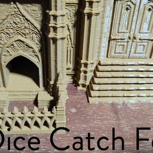 May include: A brown 3D printed miniature building with a small fence in front of it. The building has a door and a window. The fence is made of small spikes. The text "Dice Catch Fence" is printed below the fence.