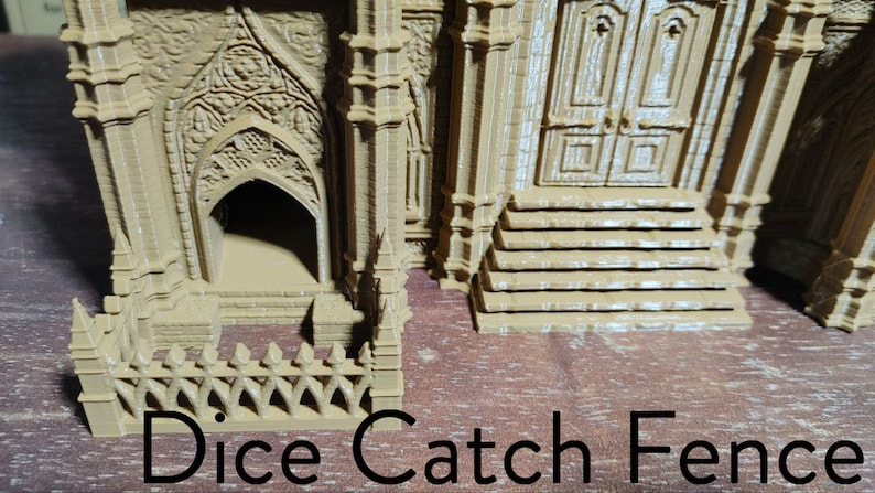 May include: A brown 3D printed miniature building with a small fence in front of it. The building has a door and a window. The fence is made of small spikes. The text "Dice Catch Fence" is printed below the fence.