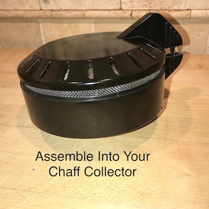 May include: A black chaff collector component, cylindrical in shape, with a mesh band and a hinged top. The text "Assemble Into Your Chaff Collector" is printed below. The item is on a wooden surface.
