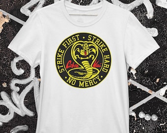 May include: White t-shirt with a black and yellow circle graphic featuring a cobra with the text "Strike First Strike Hard No Mercy" and "Cobra Kai" in red.