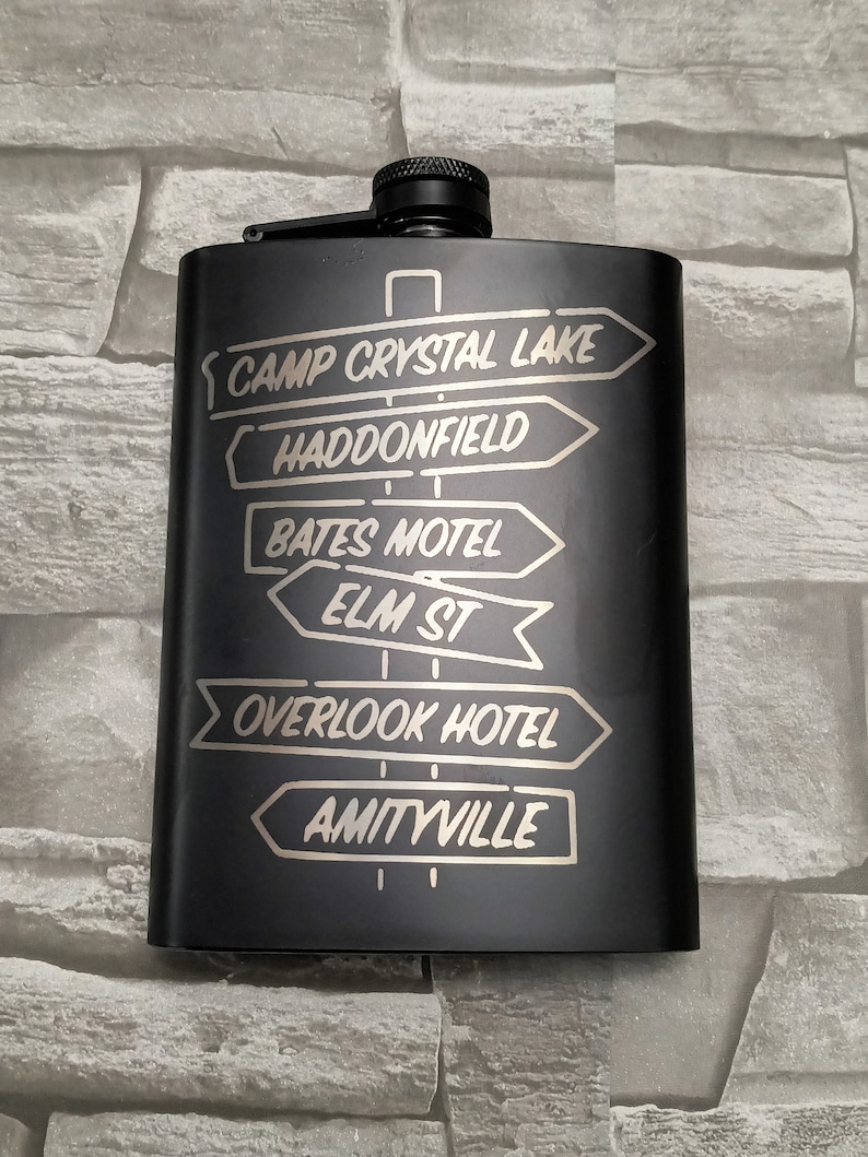 May include: Black metal flask with a silver engraved design of directional signs pointing to locations from horror movies. The signs read: Camp Crystal Lake, Haddonfield, Bates Motel, Elm St, Overlook Hotel, Amityville.