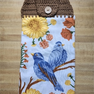May include: A hanging kitchen towel featuring a white background with a pattern of blue birds, yellow sunflowers, and orange flowers. The top section is made of brown crocheted fabric with a single light-colored wooden button for closure.