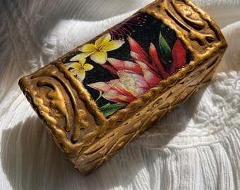 May include: A small, rectangular box with a gold finish and a floral design. The box has a raised, textured pattern on the lid and sides. The floral design features pink, yellow, and red flowers on a black background.