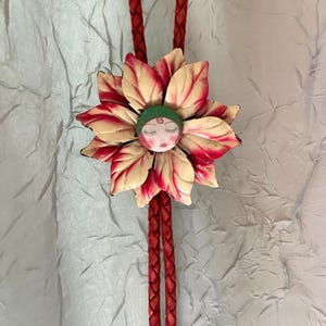 May include: A handcrafted bolo tie featuring a flower design with red and cream-colored petals. The flower has a face with closed eyes and a green cap. The tie has a braided red leather cord.