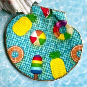 May include: A decorative shell dish featuring a blue tiled swimming pool pattern with colorful summer motifs including yellow pineapples, a rainbow popsicle, a multi-colored beach ball, a striped pool float, and orange slices with gold trim.