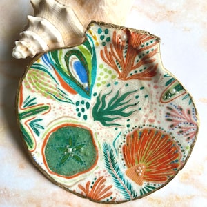 May include: A decorative scallop shell dish featuring a hand-painted pattern of orange, green, and blue sea life motifs including coral and sea urchins. The shell has a metallic gold painted rim and is embellished with small white pearl beads.