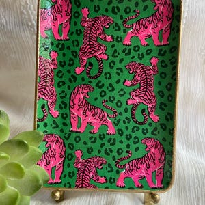 May include: A rectangular decorative tray featuring a vibrant green background with a repeating pattern of bright pink tigers and black leopard spots. The tray has a thin gold-colored rim and is supported by small gold-colored metal feet.