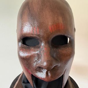 May include: A brown, full-face mask with a weathered appearance. The mask has eye openings and red accents on the forehead and around the mouth. The mask is displayed on a black surface.