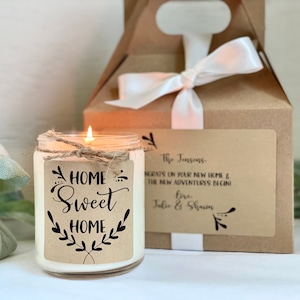 May include: A white candle in a glass jar with a brown paper label that says "HOME Sweet HOME" with black lettering and a twig design. The candle is sitting on a white surface next to a brown gift box with a white ribbon.