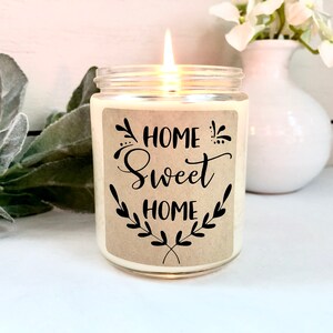 May include: A white candle in a clear glass jar with a brown label that reads "Home Sweet Home" in black script. The label has a decorative floral design.