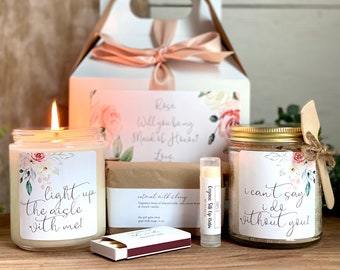 May include: A bridesmaid gift box with a floral theme. The box includes a candle with the text "light up the aisle with me!" a bar of soap, a lip balm, a jar of bath salts with the text "I can't say I do without you!" and a box of matches.