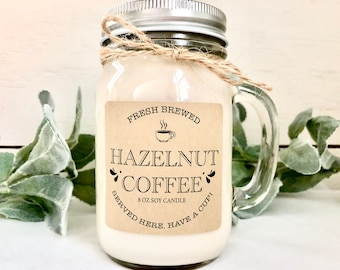 Op de afbeelding: Een glazen potkaars met een bruin etiket waarop staat "FRESH BREWED HAZELNUT COFFEE 8 OZ SOY CANDLE SERVED HERE. HAVE A CUP!"