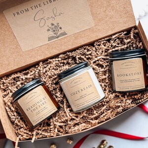 May include: A gift box filled with three candles, each with a different scent and label. The candles are labeled "Reading by the Fire", "Cozy Reads", and "Bookstore". The box is decorated with a brown paper tag that says "From the Library of Sasha".