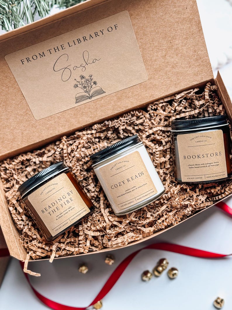 May include: A gift box filled with three candles, each with a different scent and label. The candles are labeled "Reading by the Fire", "Cozy Reads", and "Bookstore". The box is decorated with a brown paper tag that says "From the Library of Sasha".
