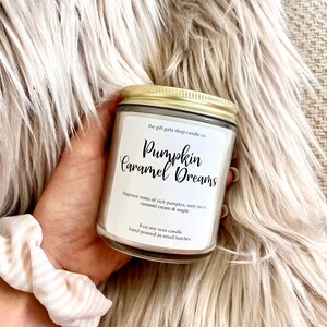 May include: A white soy wax candle with a gold lid. The label reads "the gift gala shop candle co. Pumpkin Caramel Dreams" and lists the fragrance notes as "rich pumpkin, nutty pecan, caramel cream & maple". The candle is an 8 oz soy wax candle, hand-poured in small batches.