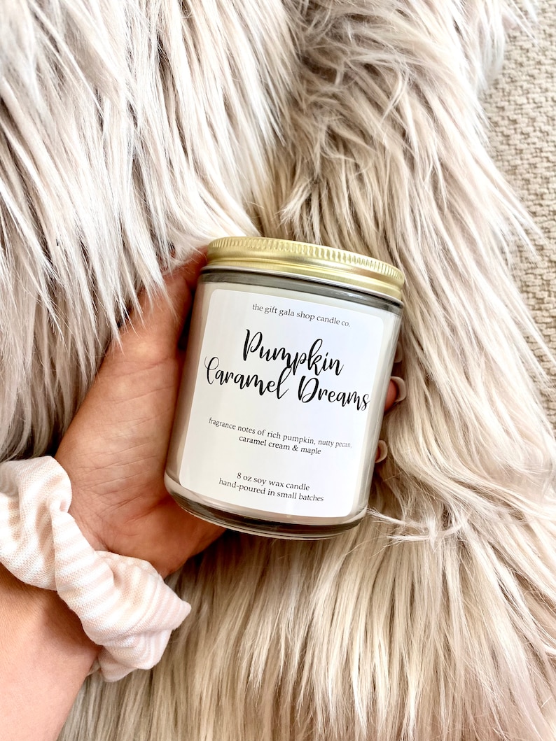 May include: A white soy wax candle with a gold lid. The label reads "the gift gala shop candle co. Pumpkin Caramel Dreams" and lists the fragrance notes as "rich pumpkin, nutty pecan, caramel cream & maple". The candle is an 8 oz soy wax candle, hand-poured in small batches.