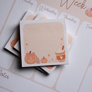 Pu&ograve; includere: Un blocchetto di foglietti adesivi quadrato con sfondo color pesca chiaro, illustrato con una zucca con fiocco, una piccola candela, una zucca pi&ugrave; piccola e una tazza di caff&egrave; con panna montata. Il blocchetto poggia su un planner settimanale con il testo This Week.