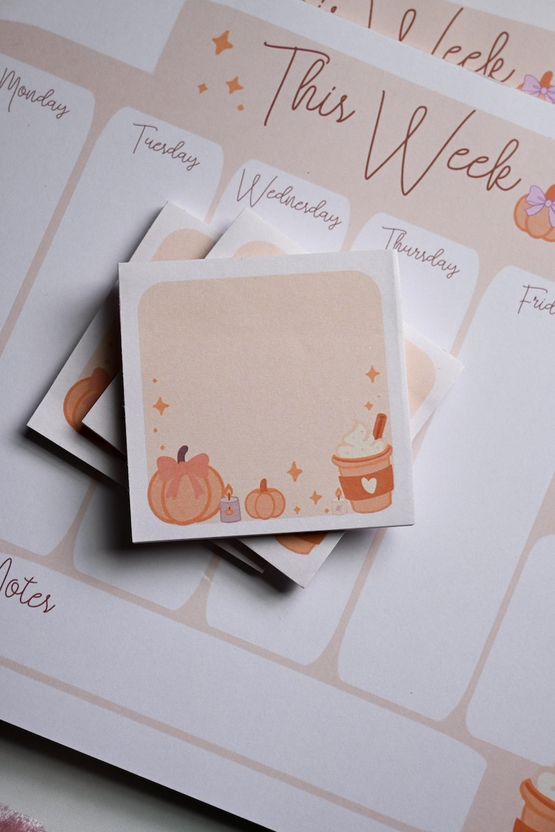 Pu&ograve; includere: Un blocchetto di foglietti adesivi quadrato con sfondo color pesca chiaro, illustrato con una zucca con fiocco, una piccola candela, una zucca pi&ugrave; piccola e una tazza di caff&egrave; con panna montata. Il blocchetto poggia su un planner settimanale con il testo This Week.