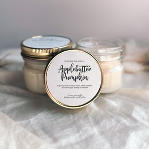 May include: Two glass jars with gold lids filled with a white candle. The jar on the left has a label that reads "the gift gable shop candle co. Applebutter Pumpkin" with the text "Fragrance notes of buttery vanilla, baked pumpkin, cinnamon apple, nutmeg & cinnamon. 3 oz soy wax candle hand-poured in small batches."