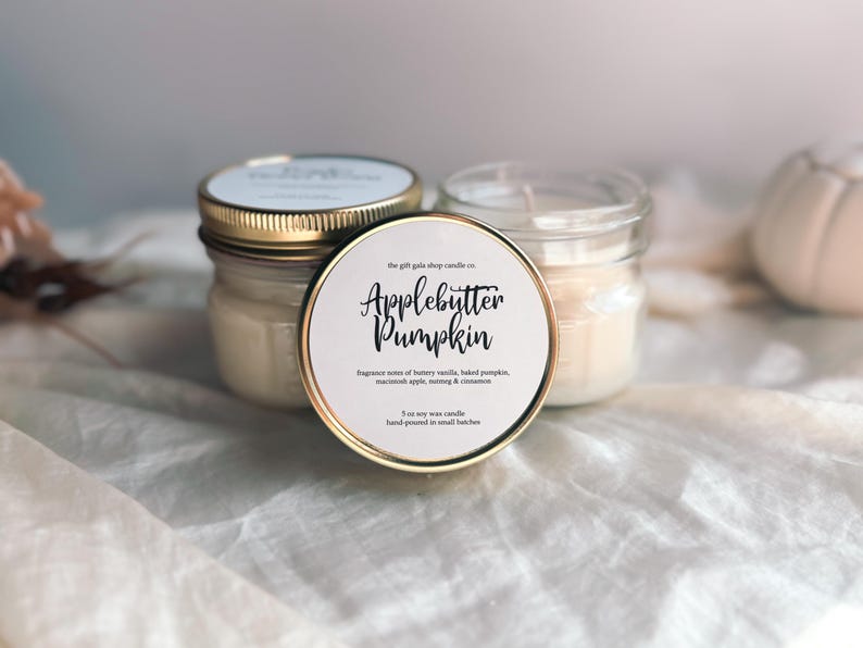 May include: Two glass jars with gold lids filled with a white candle. The jar on the left has a label that reads "the gift gable shop candle co. Applebutter Pumpkin" with the text "Fragrance notes of buttery vanilla, baked pumpkin, cinnamon apple, nutmeg & cinnamon. 3 oz soy wax candle hand-poured in small batches."