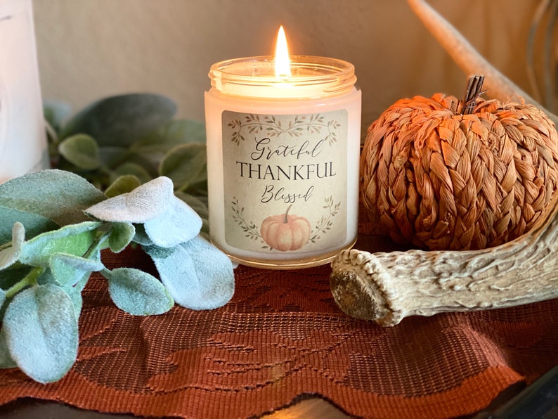 May include: A lit candle in a glass jar with a label that reads "Grateful, Thankful, Blessed" with a pumpkin illustration. A woven pumpkin decoration and green foliage are also visible, creating a fall theme.