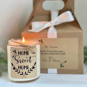 May include: A white candle in a glass jar with a brown paper label that says "Home Sweet Home" with a twig and leaf design. The candle is lit and the flame is visible. The candle is sitting on a white surface next to a brown box with a white ribbon.
