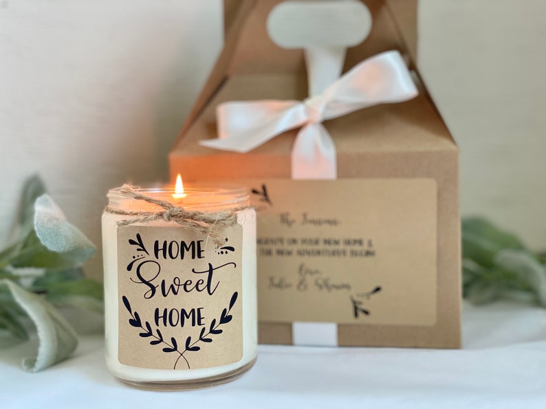 May include: A white candle in a glass jar with a brown paper label that says "Home Sweet Home" with a twig and leaf design. The candle is lit and the flame is visible. The candle is sitting on a white surface next to a brown box with a white ribbon.
