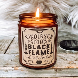 May include: A brown glass candle jar with a black and white label that reads "Sanderson Sisters Black Flame Candle Company Est. 1692". The candle is lit and the flame is visible.