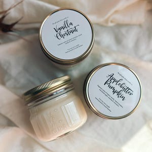May include: Two glass jars with gold lids containing soy wax candles. The labels on the jars read "Vanilla Chestnut" and "Applebutter Pumpkin". The labels list the fragrance notes and state that the candles are hand-poured in small batches.