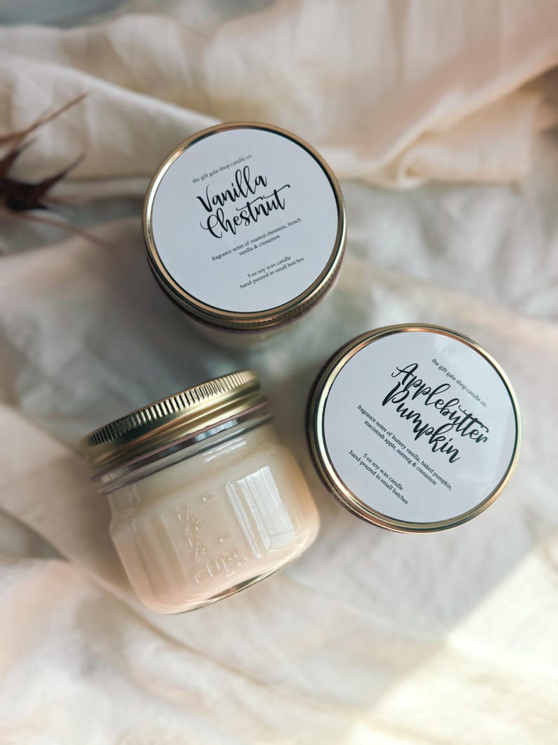 May include: Two glass jars with gold lids containing soy wax candles. The labels on the jars read "Vanilla Chestnut" and "Applebutter Pumpkin". The labels list the fragrance notes and state that the candles are hand-poured in small batches.