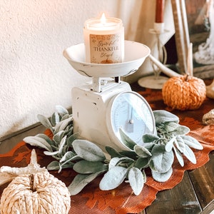 May include: A lit candle in a glass jar sits on a vintage white scale. The candle label reads "Grateful, Thankful, Blessed" with a pumpkin illustration. The scale is surrounded by faux greenery and rests on an orange lace runner, with decorative pumpkins.