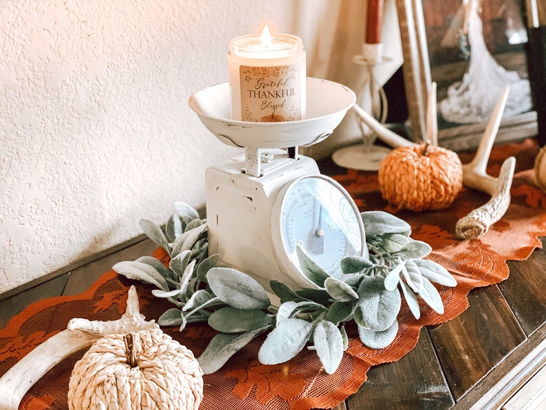 May include: A lit candle in a glass jar sits on a vintage white scale. The candle label reads "Grateful, Thankful, Blessed" with a pumpkin illustration. The scale is surrounded by faux greenery and rests on an orange lace runner, with decorative pumpkins.