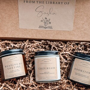 May include: Three brown glass candle jars with black lids and white labels. The labels read "Reading by the Fire", "Cozy Reads", and "Bookstore". The candles are in a brown cardboard box with the words "From the Library of Sasha" on the lid.
