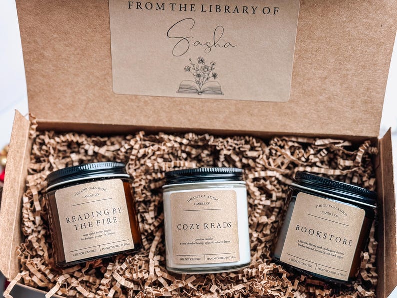 May include: Three brown glass candle jars with black lids and white labels. The labels read "Reading by the Fire", "Cozy Reads", and "Bookstore". The candles are in a brown cardboard box with the words "From the Library of Sasha" on the lid.