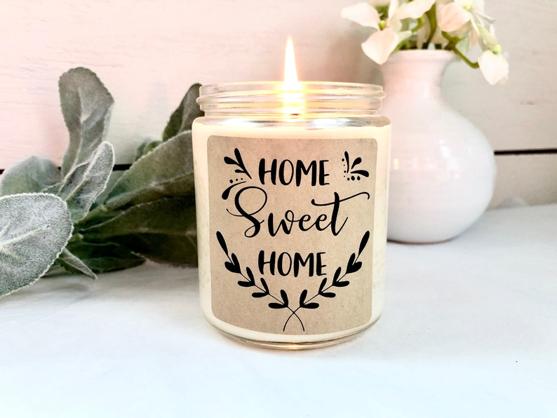May include: A white candle in a clear glass jar with a brown label that reads "Home Sweet Home" in black script. The label has a decorative floral design.
