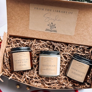 May include: A gift box filled with brown shredded paper and three candles. The candles are labeled "Reading by the Fire", "Cozy Reads", and "Bookstore". The box has a label that says "From the Library of Sasha".
